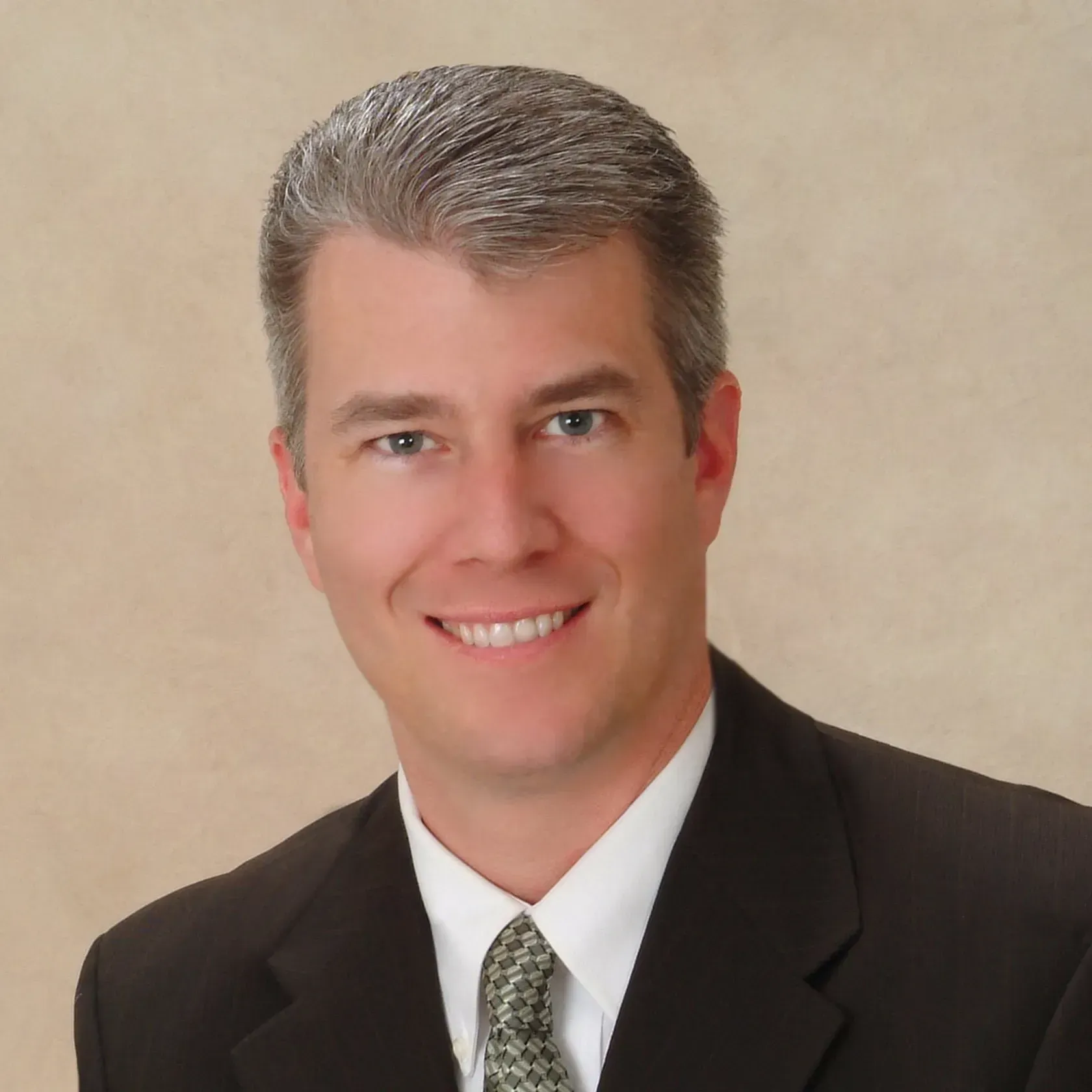 Professional headshot of a smiling person in a dark suit and tie against a beige background