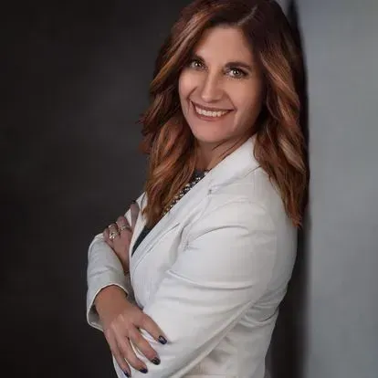 Smiling person in a white blazer posing with folded arms against a dark studio backdrop