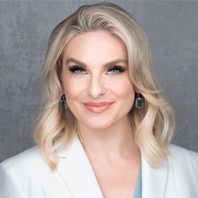 Studio headshot of a blonde person in a white blazer, smiling against a gray background
