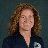 Smiling person with curly hair in a dark blue shirt against a plain dark background