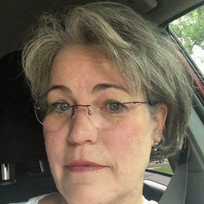 Close-up of a person with short gray hair and glasses sitting in a car, looking at the camera.
