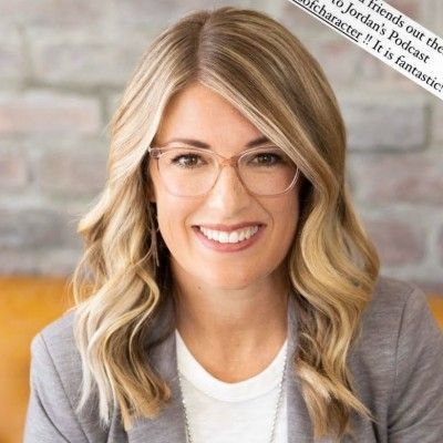 Smiling person with blonde hair and glasses in a gray blazer, against a softly blurred background