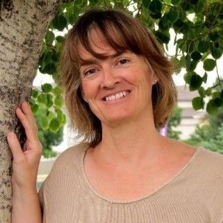 Smiling person leaning against a tree, wearing a beige top with leafy greenery in the background