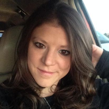 Woman with long dark hair sitting in a car, smiling softly, hand behind her head.