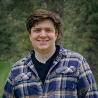 Smiling person in a blue plaid shirt standing outdoors with blurred greenery behind them