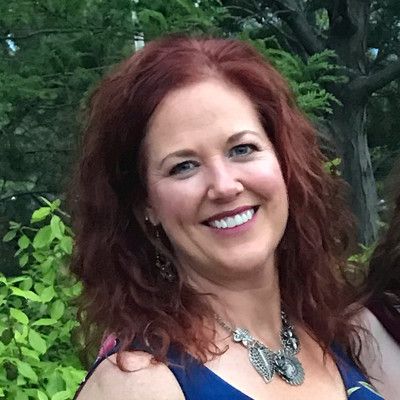 Smiling woman with red hair wearing a necklace, standing outdoors near green foliage.