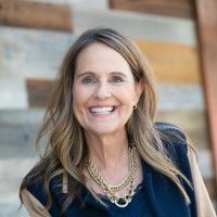 Smiling woman in a navy top and layered necklace against a wooden wall.