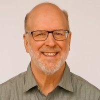 Smiling bald man with glasses and a gray beard in a gray shirt on a plain background