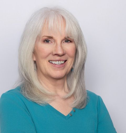 Portrait of a smiling woman with long white hair wearing a turquoise top against a light background