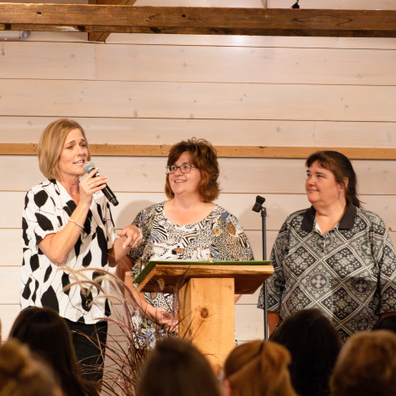 Three women standing on stage, one speaking into a microphone beside a wooden lectern.