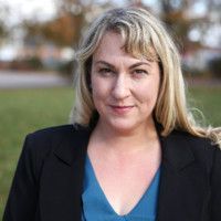 Portrait of a blonde woman in a black blazer and blue top outdoors, with a blurred background