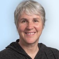 Smiling person with short gray hair wearing a dark hoodie against a light blue background