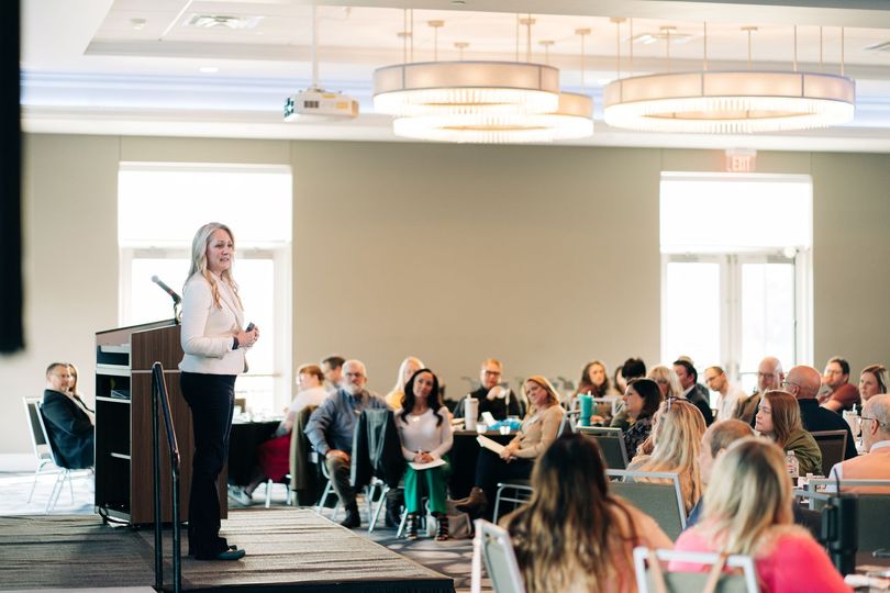 Presenter speaking at podium to seated audience in a bright conference room