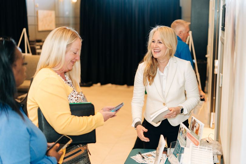 Two women chatting at a booth, one holding a phone and the other smiling in a white blouse.