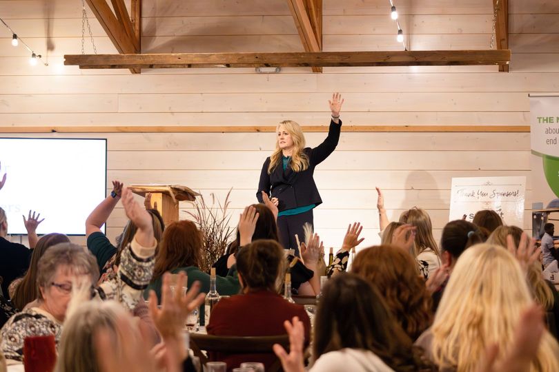Speaker on stage addressing a standing crowd in a wooden hall, hands raised in participation