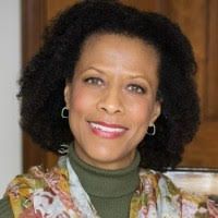 Smiling woman in a green turtleneck and floral top, seated indoors against a warm wood background.
