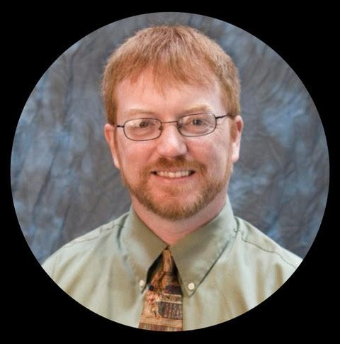 Portrait of a smiling person with glasses, red hair, and a patterned tie against a blue-gray studio backdrop