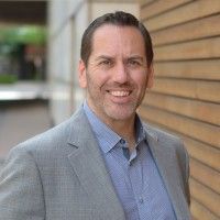 Smiling man in a gray blazer and blue shirt standing outdoors beside a wooden wall