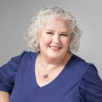 Smiling person with curly gray hair wearing a blue top on a light gray background