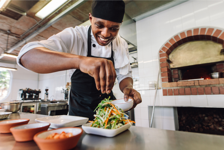 Chef plating a colorful salad in a bright kitchen near a brick oven
