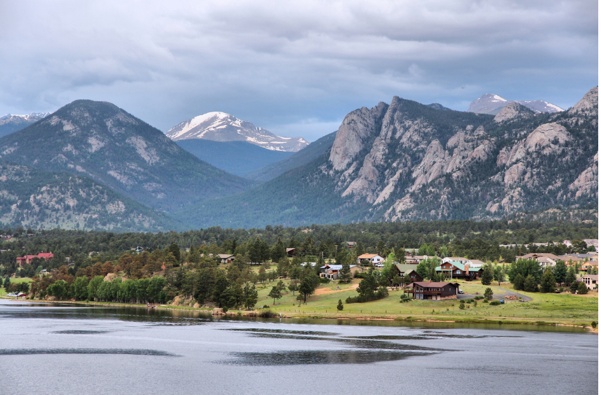 Mountain lake with a small town, pine trees, and snow-capped peaks under a cloudy sky