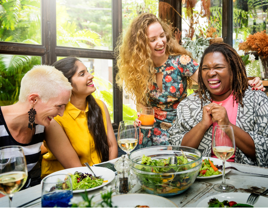 Friends laughing and sharing a meal at a sunlit dining table indoors