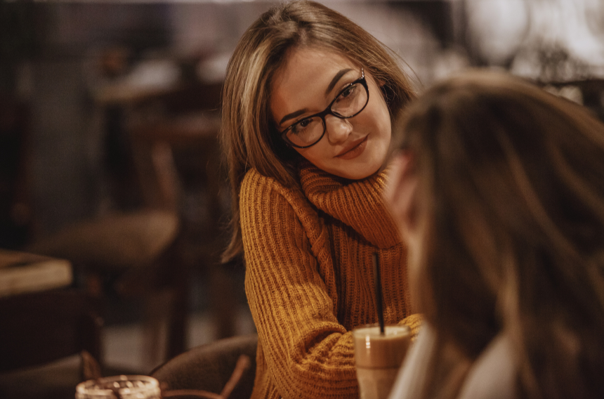 Woman in a rust sweater and glasses leaning over a café table, smiling at someone off-camera