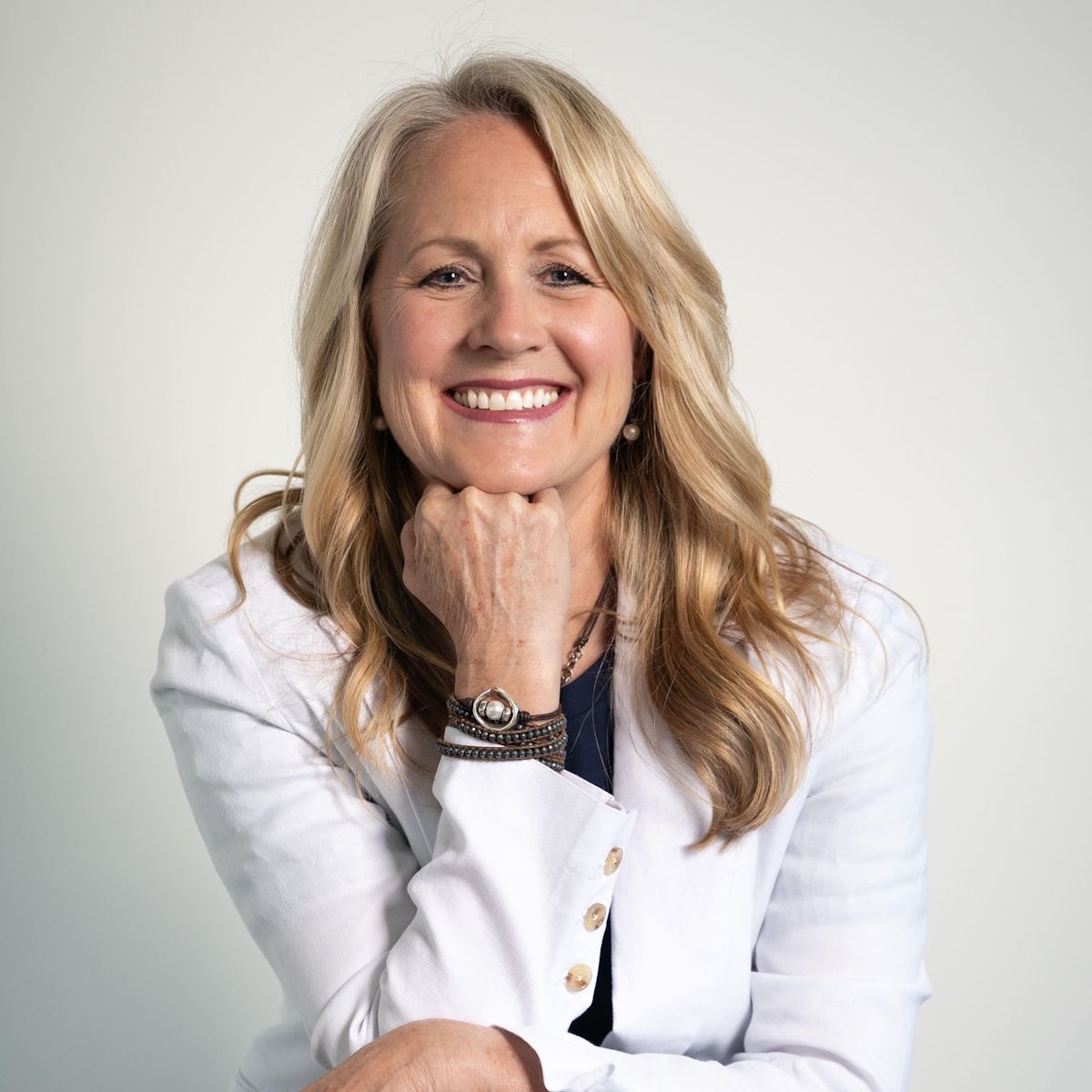 Smiling blonde woman in white blazer, resting chin on hand against a light gray background