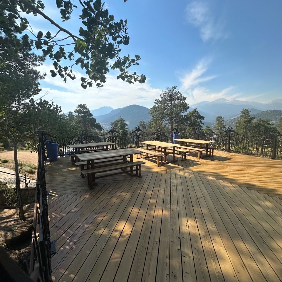 Wooden deck with picnic tables overlooking trees, mountains, and a blue sky at sunrise.