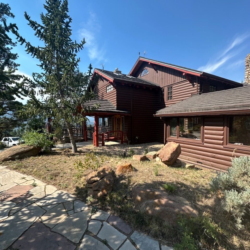 Rustic log cabin exterior with red-trimmed porch, stone path, and pine trees under a blue sky