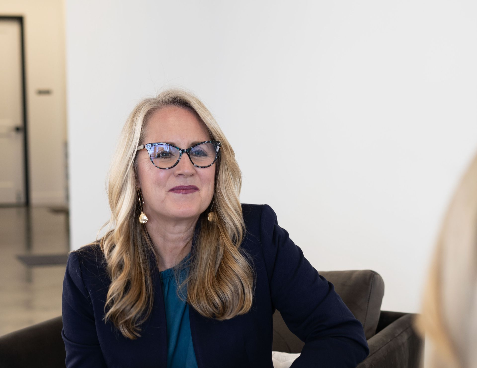 Woman with glasses speaking in a bright office setting, wearing a navy blazer and teal top.