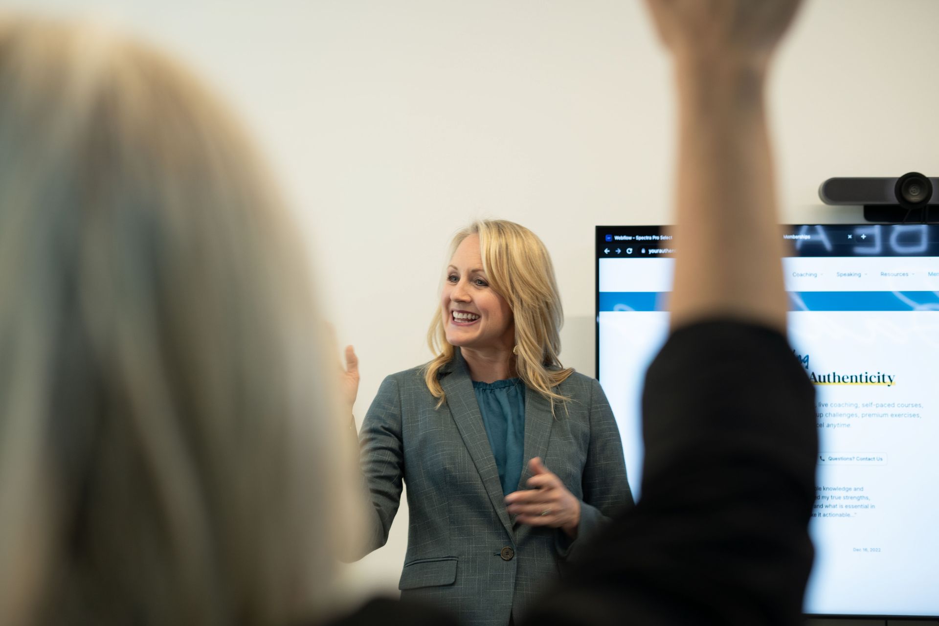 Presenter speaking in a meeting room beside a screen while attendees raise their hands