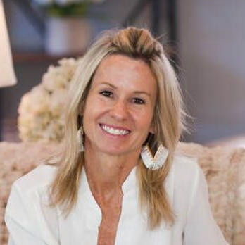 Smiling blonde woman in a white blouse, seated indoors on a light-colored couch