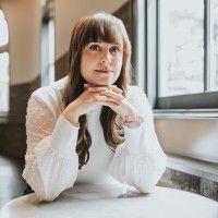 Woman with long brown hair rests her chin on folded hands at a table beside a bright window.