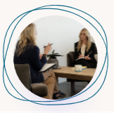 Two women in a meeting, seated across a table in a bright office lounge.