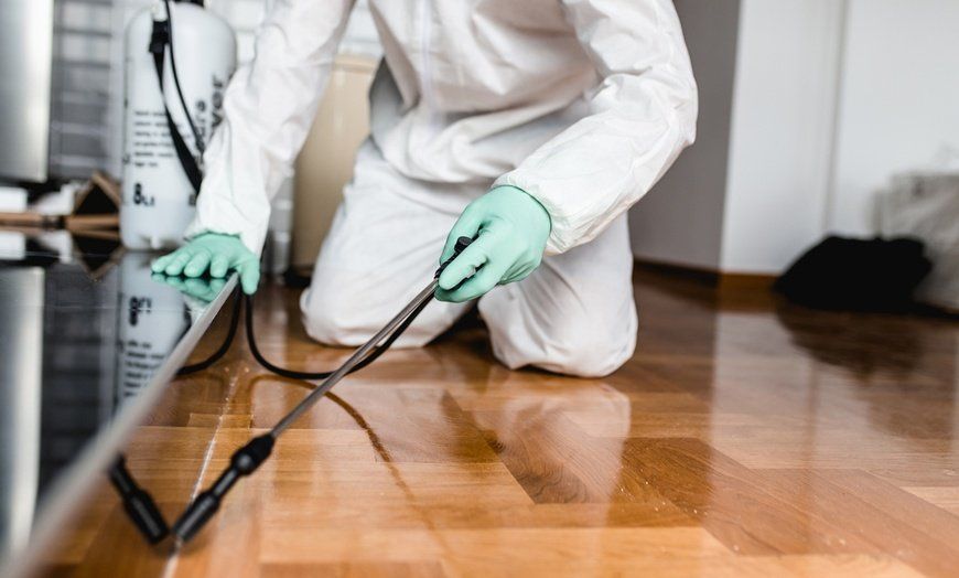 A man in a protective suit is spraying a wooden floor with a sprayer.