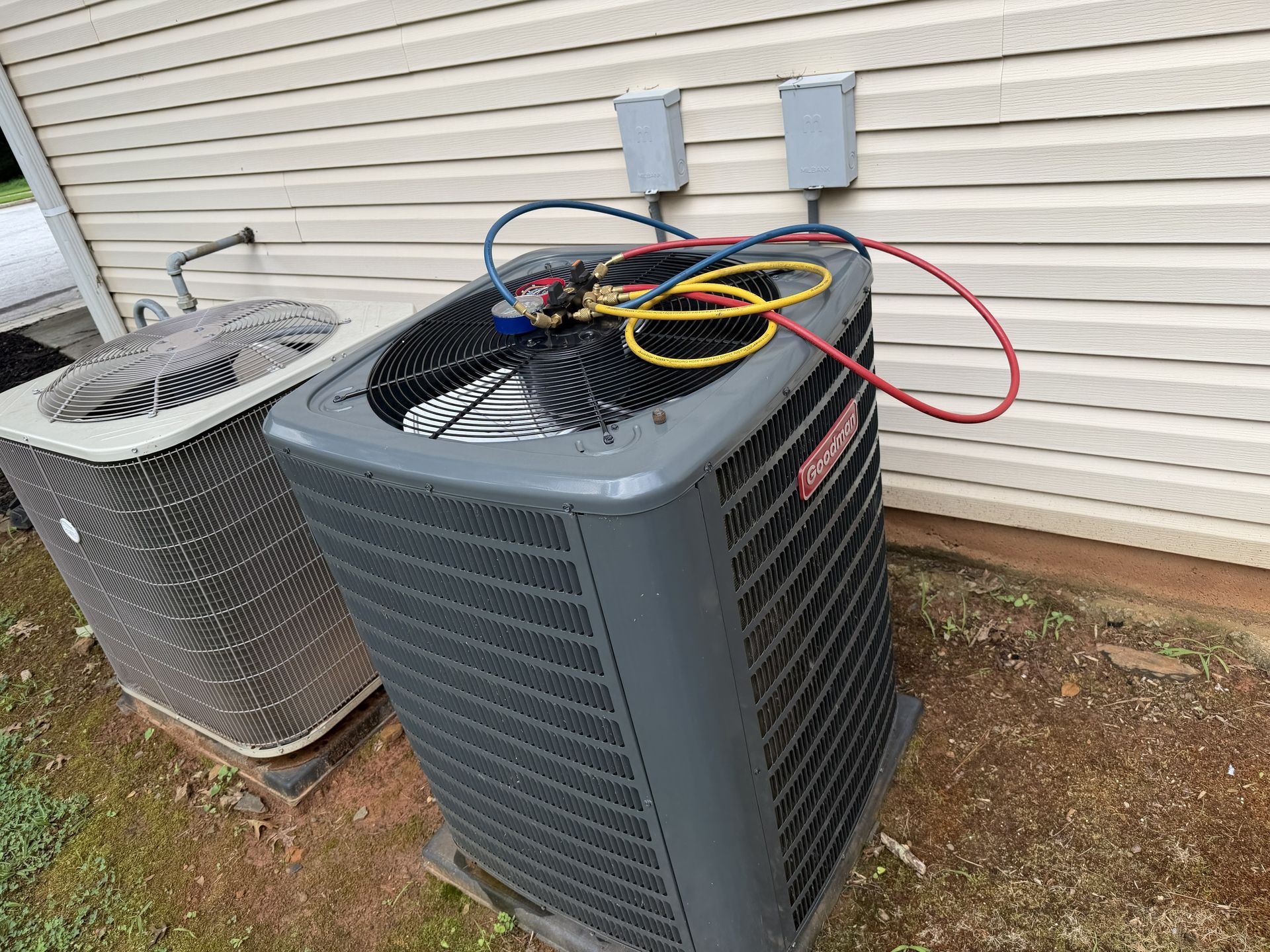 Two outdoor HVAC units beside a house, with gauges and hoses on the right unit.