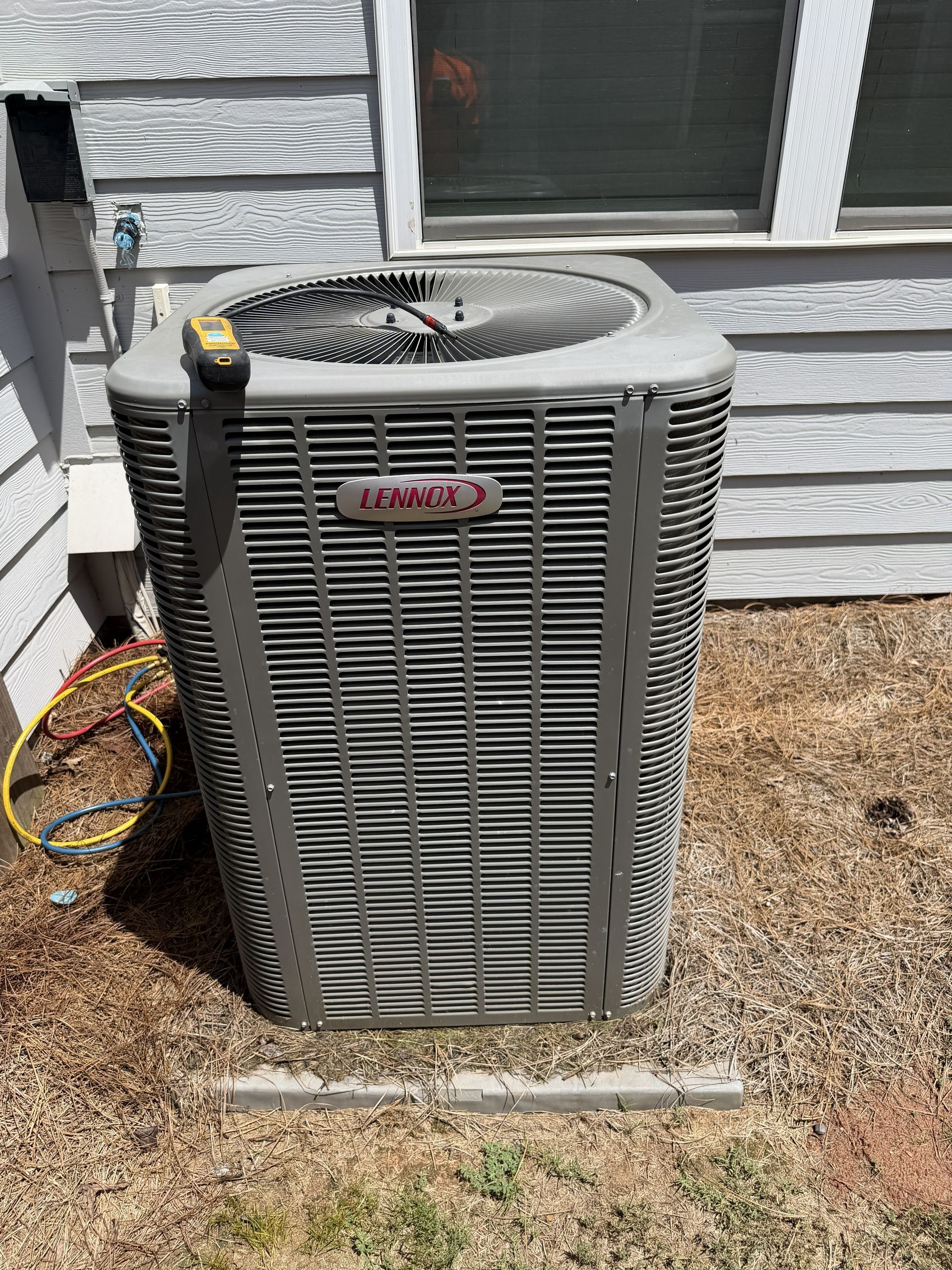 Outdoor air conditioner unit beside a house wall, with hoses and wires attached.