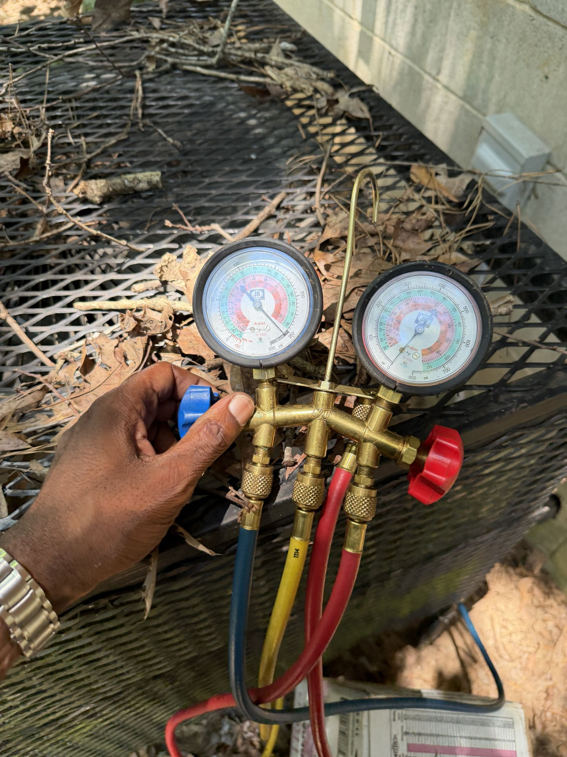 Technician’s hand holding HVAC manifold gauges with colored hoses on an outdoor AC unit.