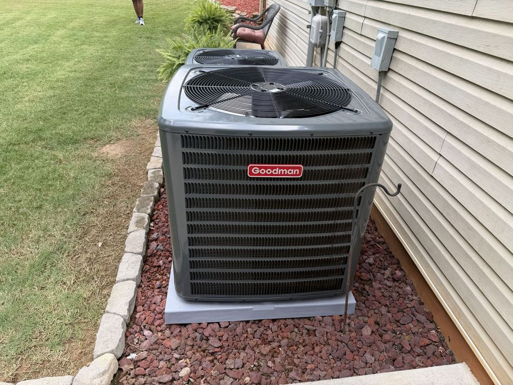 Outdoor air conditioner unit beside a house with a fan cover on top, sitting in a rock bed along the siding.