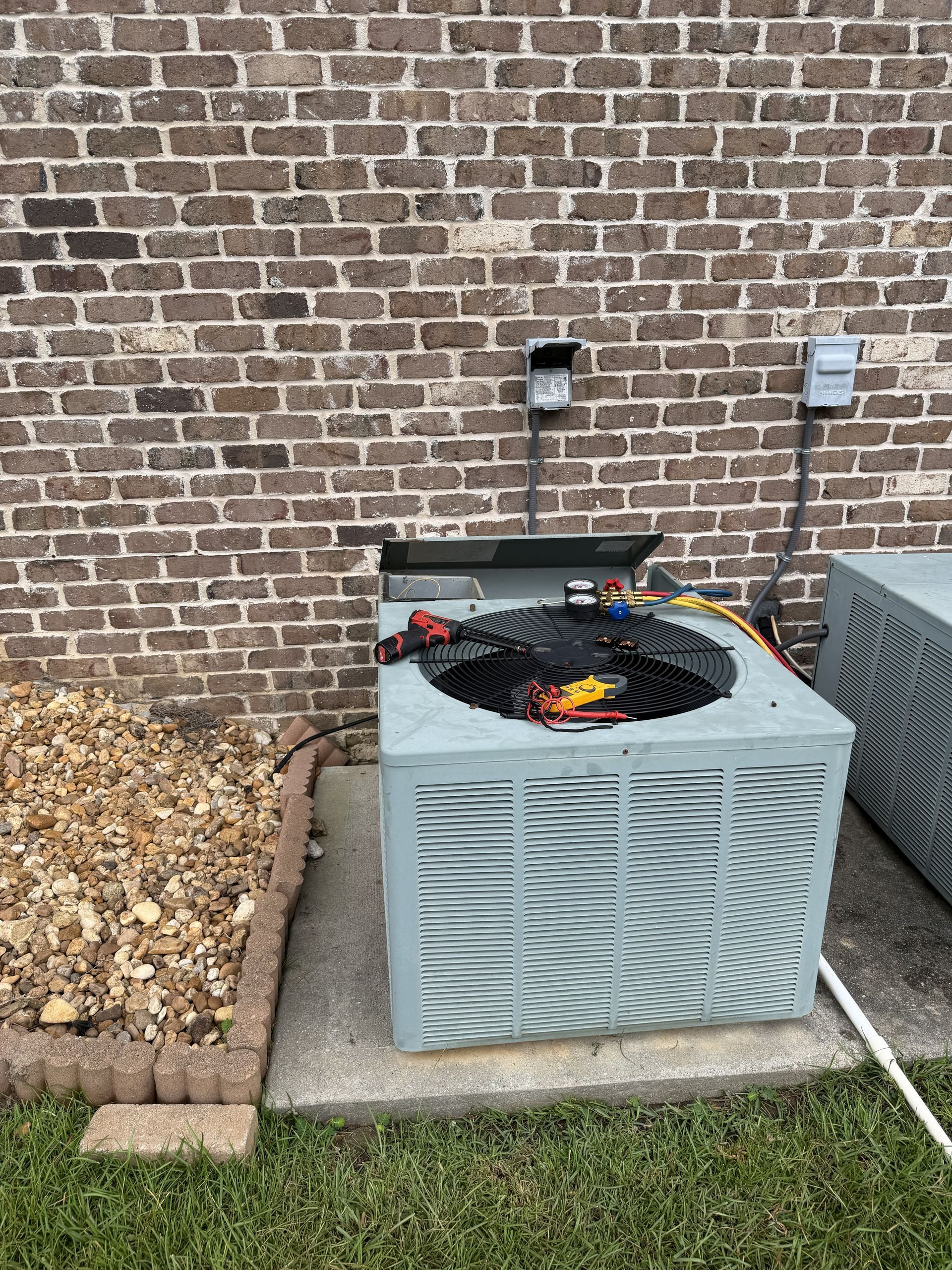 Outdoor HVAC unit beside brick wall with debris on top and a white hose on the ground