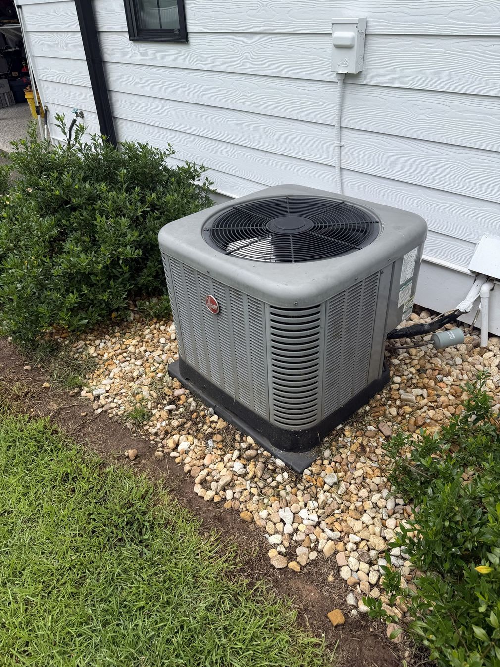 Outdoor central air conditioner unit beside a white house wall and shrubs.