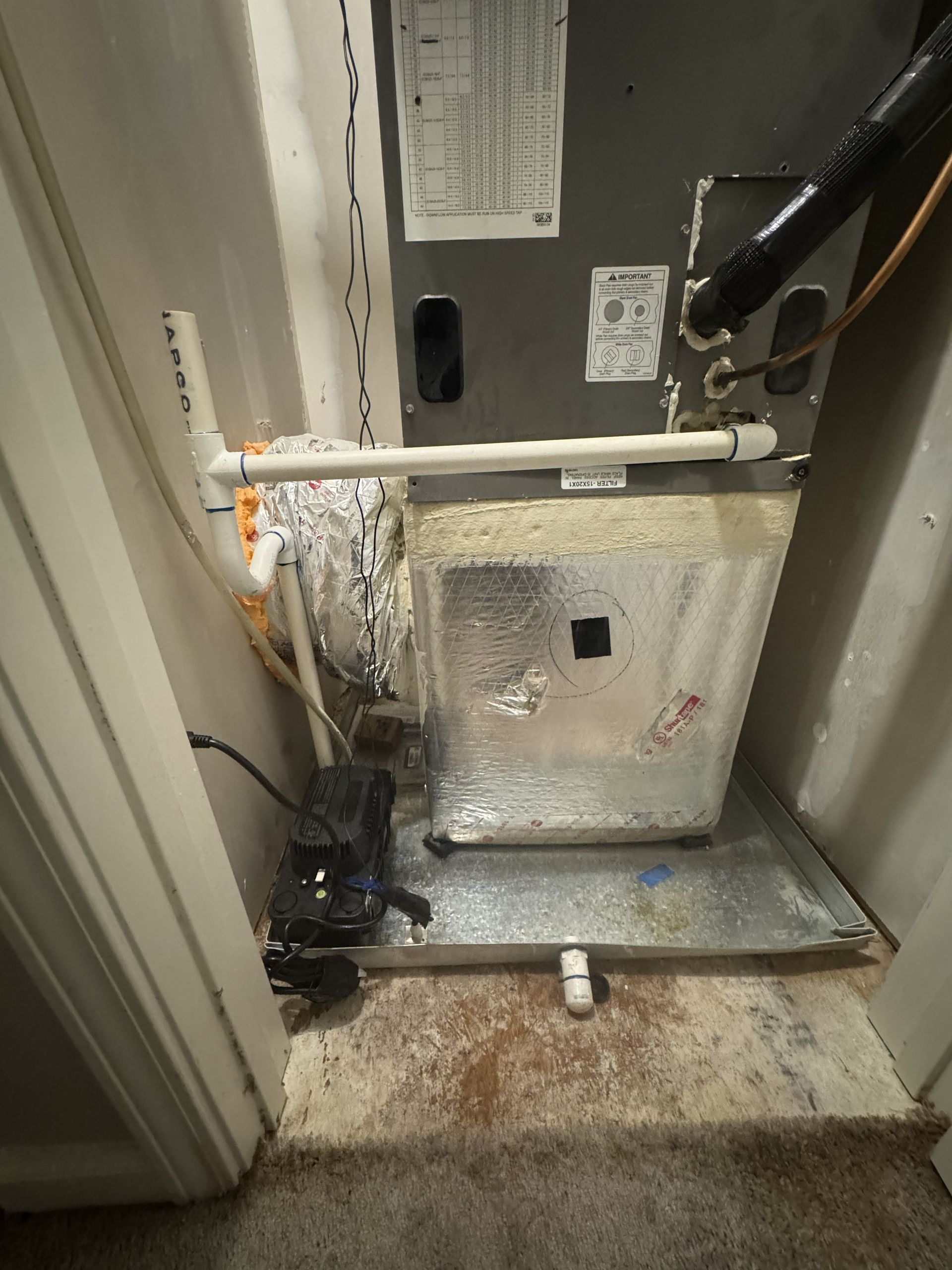 HVAC furnace unit in a utility closet with exposed pipes, wiring, and a dirty floor panel