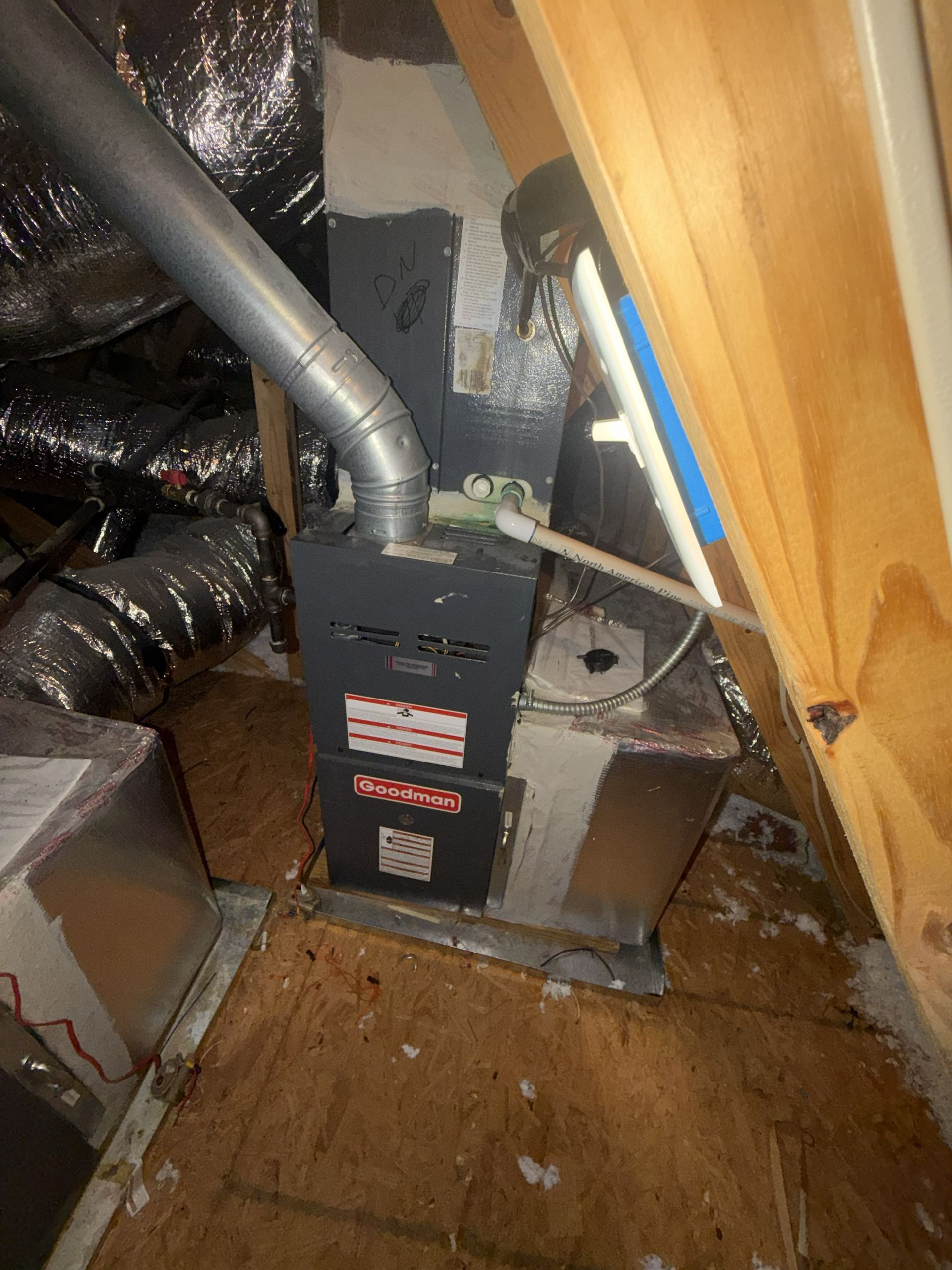 Furnace and ductwork in a cramped attic space with exposed wood framing and insulation.