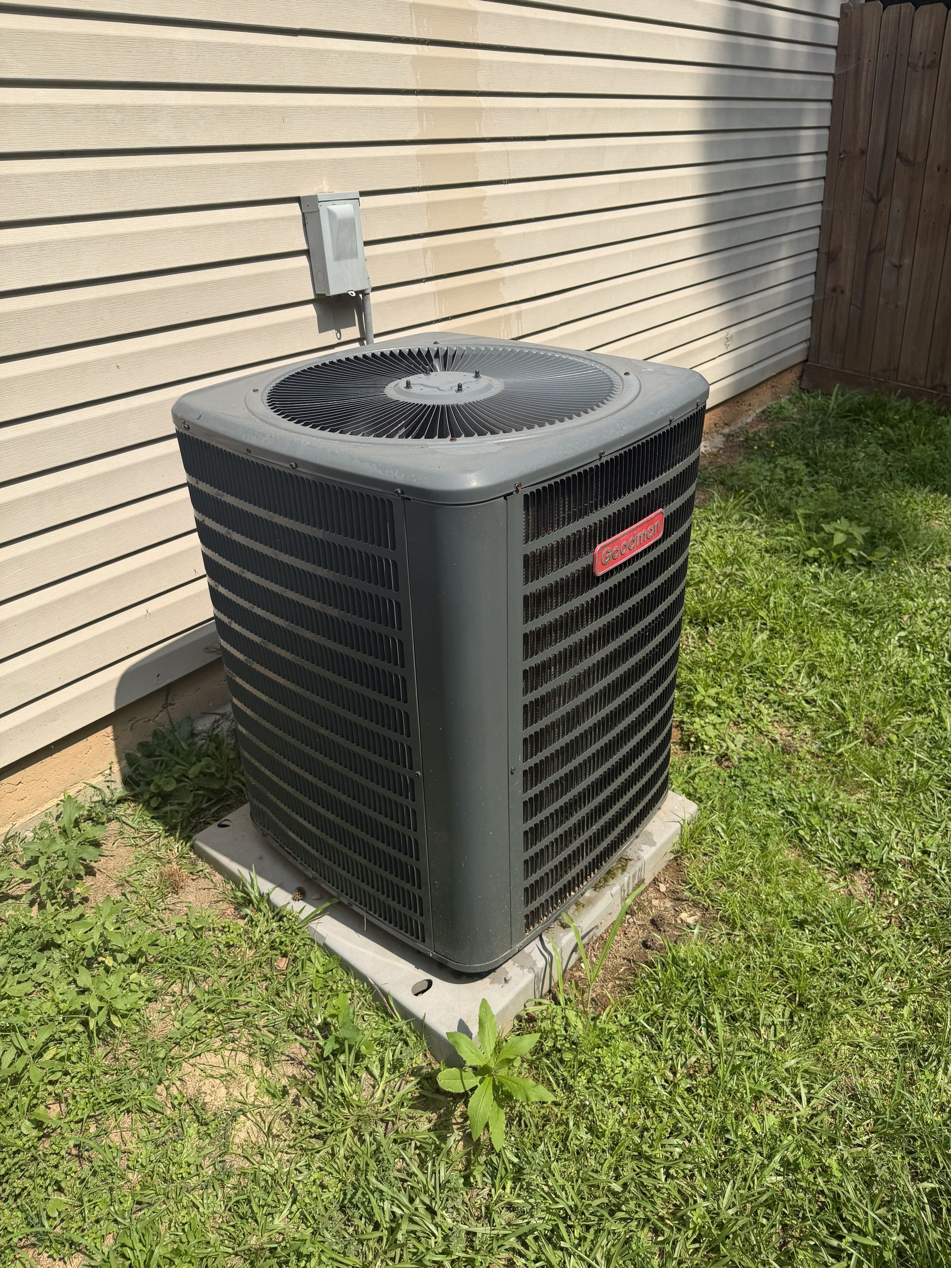 Outdoor air conditioner unit beside a house wall on a concrete pad, with grass around it.