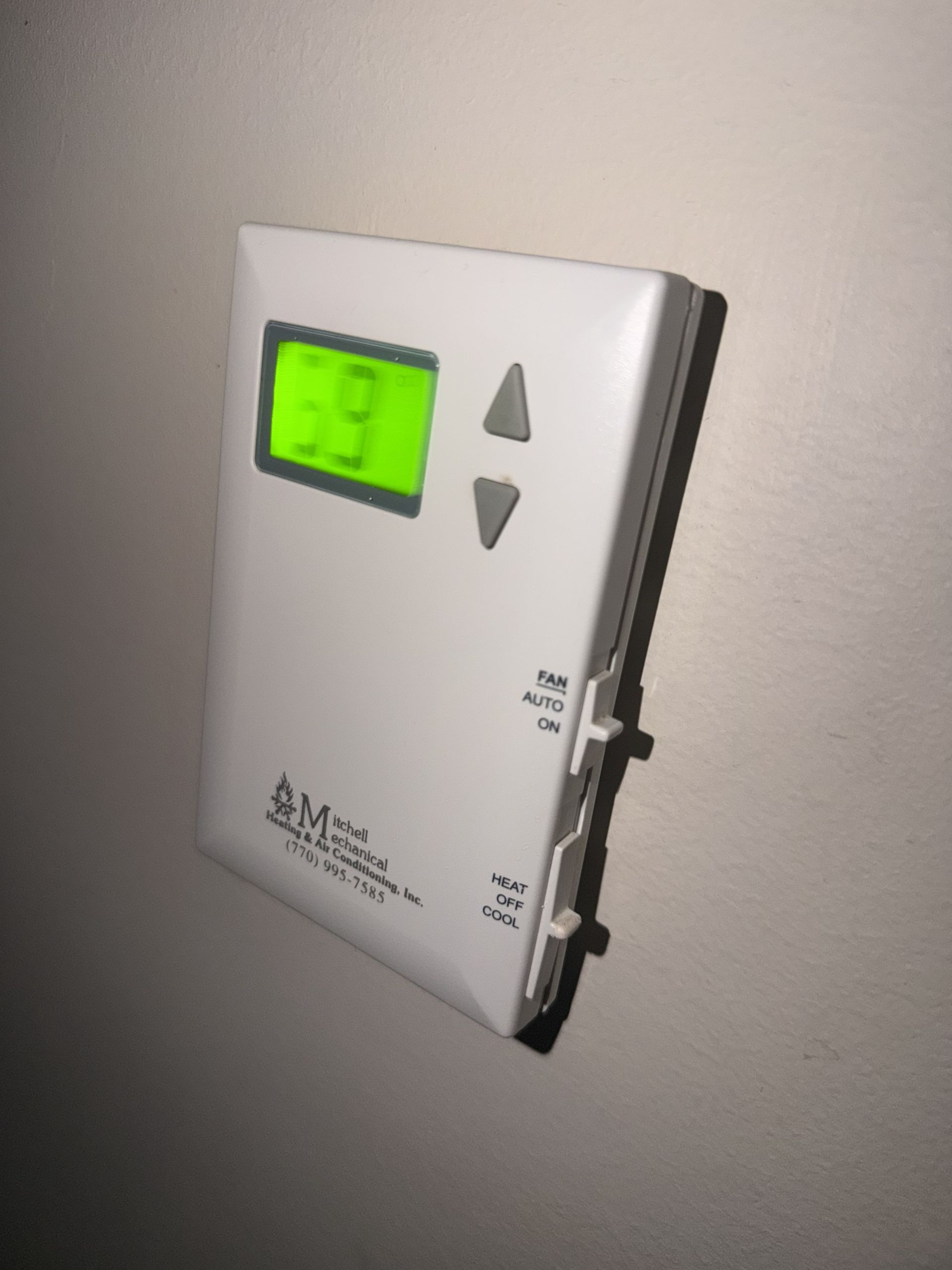 Wall-mounted thermostat with green digital display and up/down arrow buttons on a white panel.