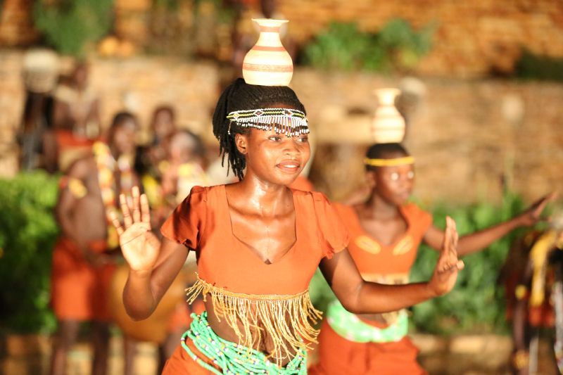 Woman in orange top balances a pot on her head while dancing with others outdoors.