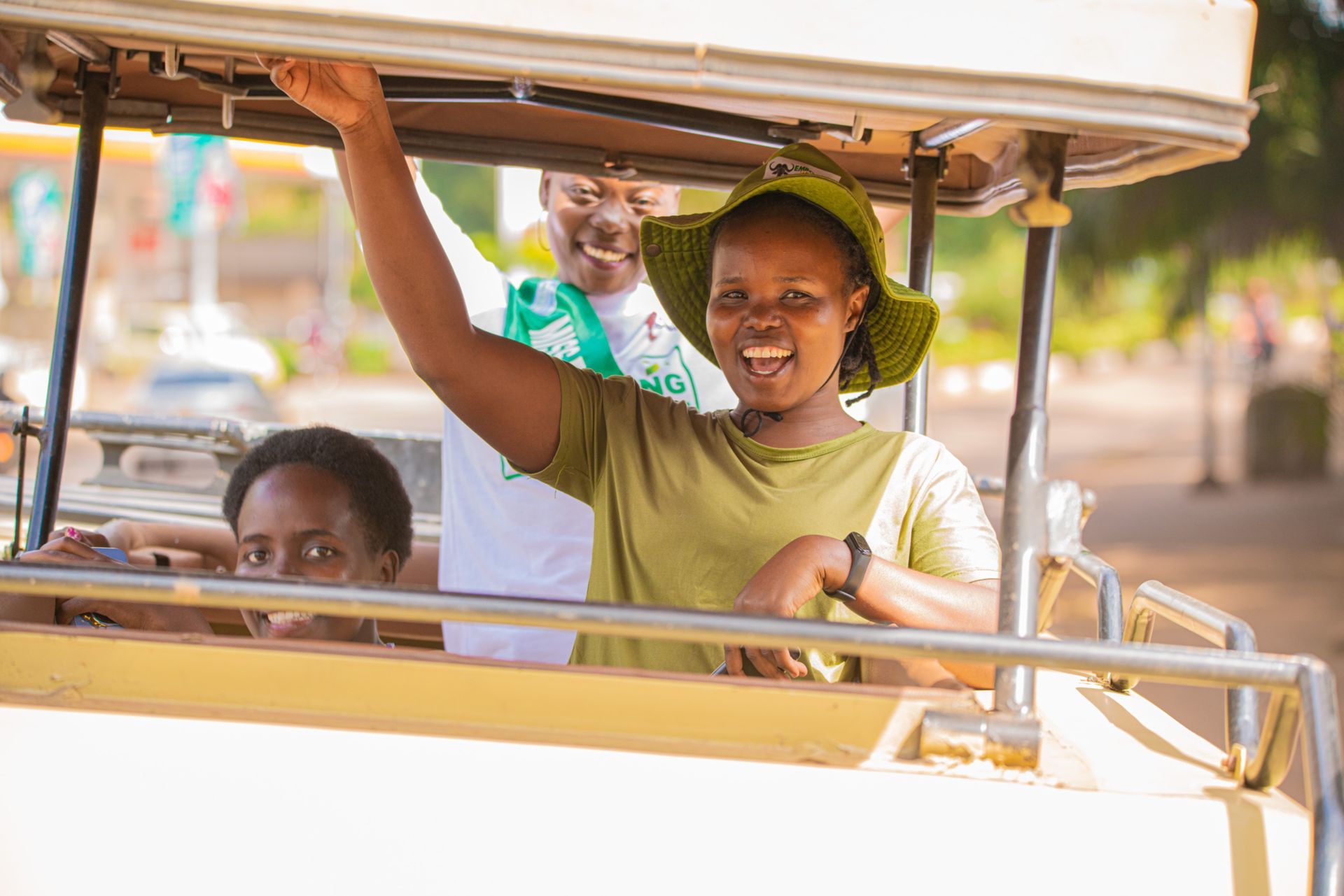 Three people smiling, waving from a light-colored vehicle under a bright sky.