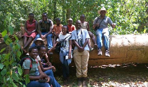 Group of people sitting and standing on a fallen tree in a forest, smiling, some holding binoculars.