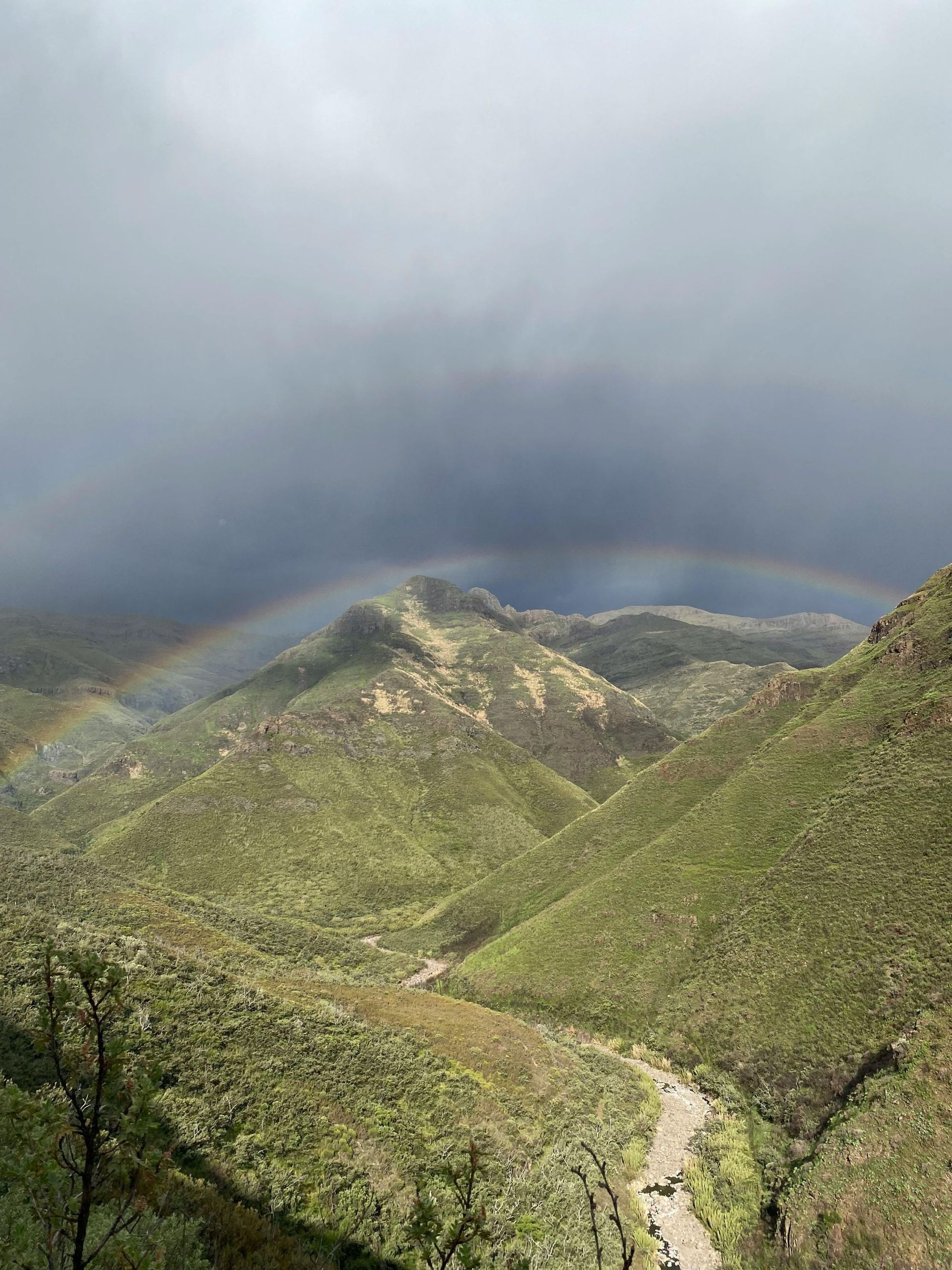 Rainbow arcs over green, mountainous terrain with a dark, stormy sky in the background.